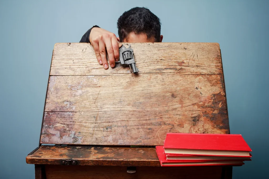 Person hiding behind a school desk with a handgun, representing school violence and the need for school security planning